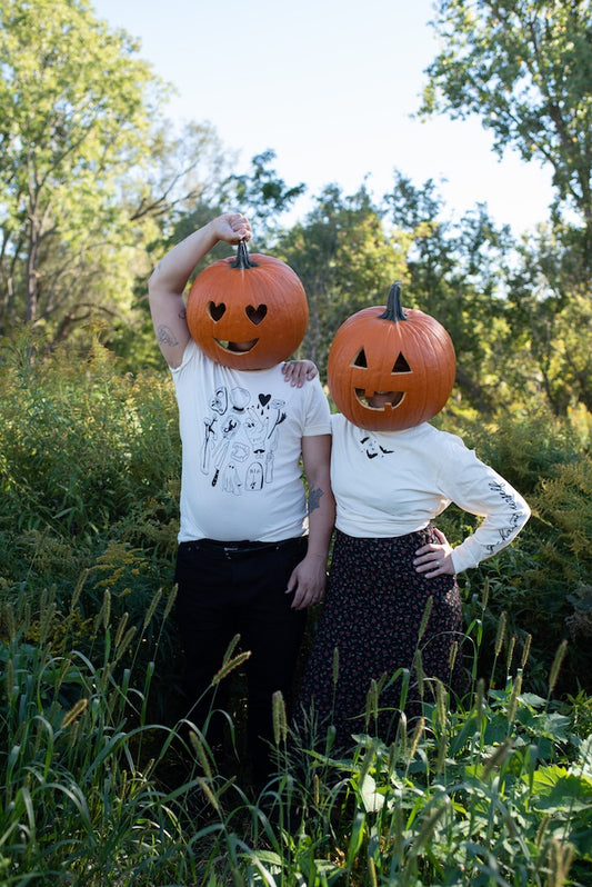 two people standing in a lush green field wearing carved pumpkins on their heads. the people are wearing a short sleeved tshirt with halloween drawings and a long sleeve shirt that says go haunt urself down the sleeves