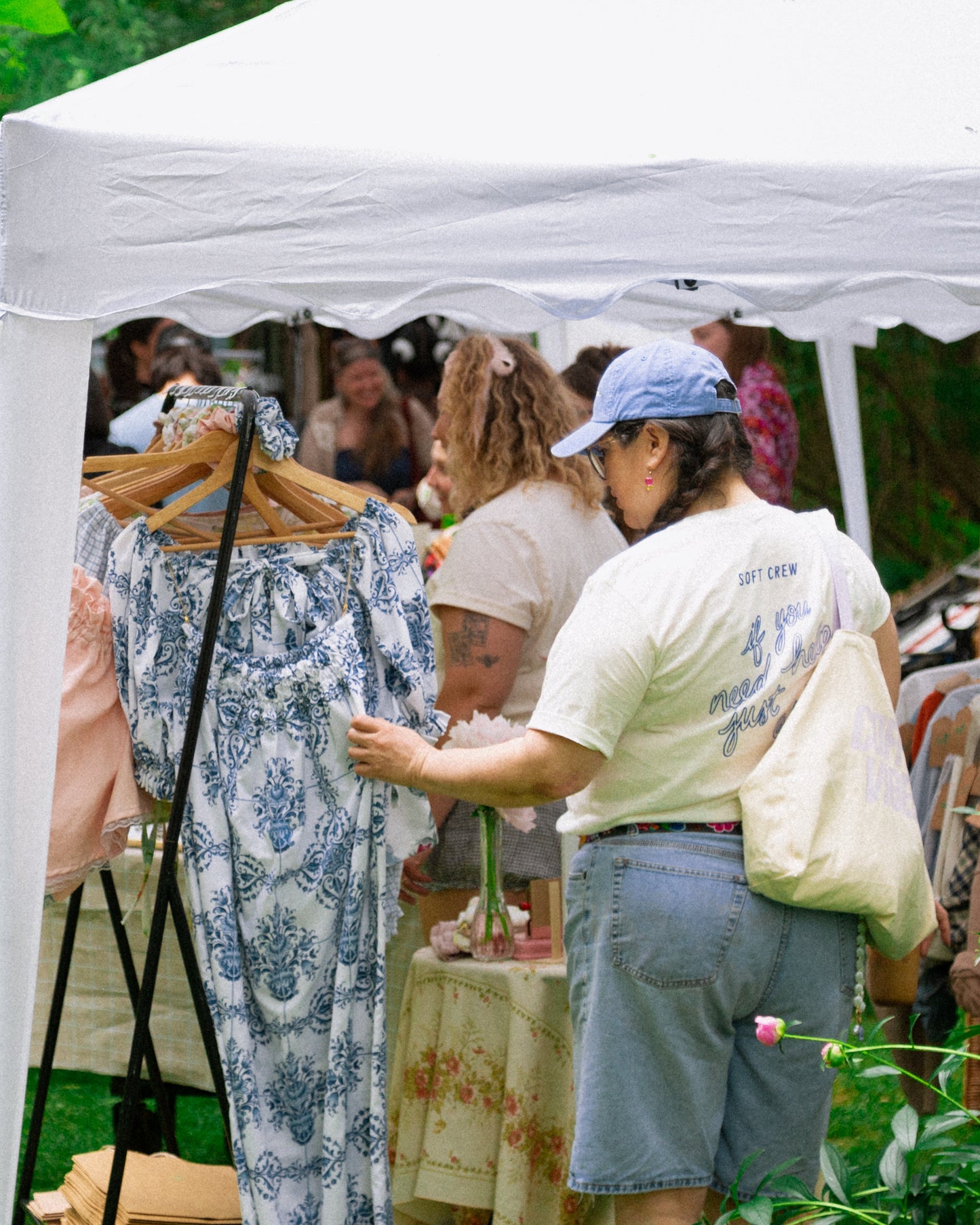 People shopping at an outdoor flea market under a white tent with clothing on display.