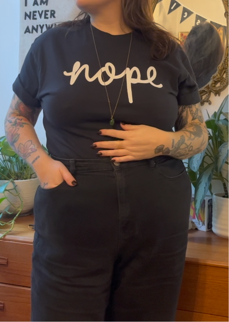 A person wearing a black graphic t-shirt with the word 'nope' printed in white script across the chest, paired with black jeans. The setting is a cozy indoor room with wooden furniture and potted plants in the background.