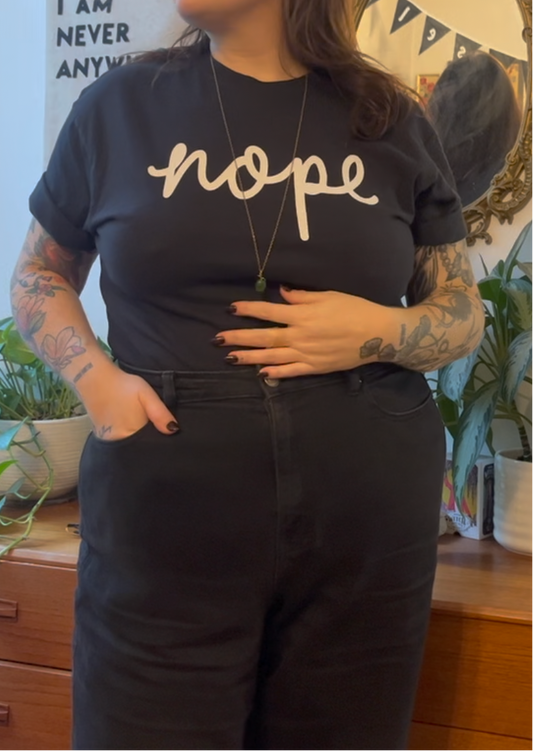 A person wearing a black graphic t-shirt with the word 'nope' printed in white script across the chest, paired with black jeans. The setting is a cozy indoor room with wooden furniture and potted plants in the background.